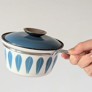 Puede incluir: Una cacerola de esmalte blanco con un patrón de hojas azules y una tapa de esmalte azul. La cacerola tiene un asa plateada.