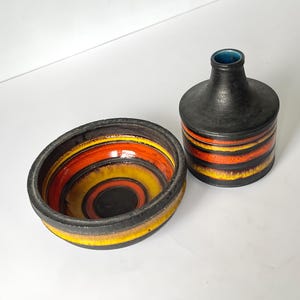 Op de afbeelding: Een keramische kom en fles. De kom heeft concentrische ringen van geel, oranje en zwart. De fles heeft oranje strepen en een blauwe binnenkant. Afmetingen in cm.