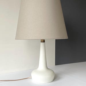 May include: A white ceramic table lamp with a beige fabric lampshade. The lamp has a simple, elegant design with a rounded base and a tall, slender neck.