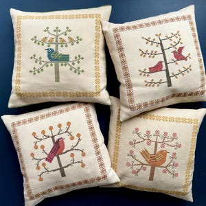 May include: Four square throw pillows with embroidered bird and tree designs. Each pillow features a different bird and tree pattern in shades of green, red, orange, and brown on a cream background. The pillows have a decorative border.