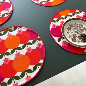 Op de afbeelding: Ronde stoffen placemats met een herhalend patroon van oranje, rode en magenta cirkels met kleine roze bloemen en donkergroene stelen op een witte achtergrond. Op één placemat staat een wit bord met een bruin bloemmotief en bestek.