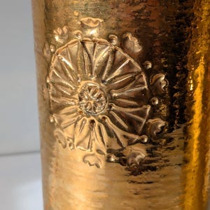 May include: Gold-toned ceramic vase with a raised embossed floral design. The vase has a textured surface and a cylindrical shape.