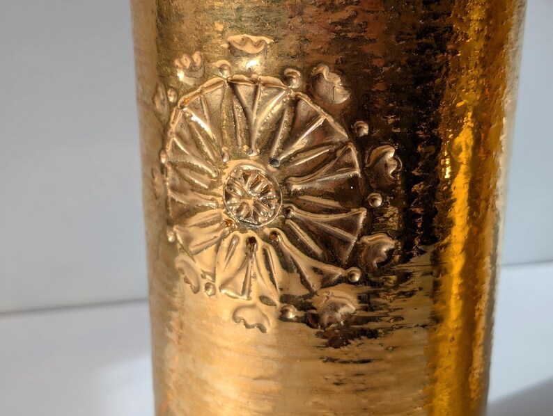 May include: Gold-toned ceramic vase with a raised embossed floral design. The vase has a textured surface and a cylindrical shape.