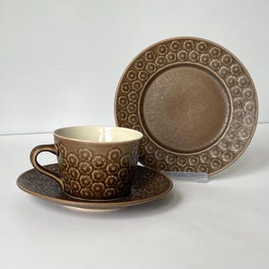 以下が含まれることがあります： 茶碗、ソーサー、お皿のセット。茶碗とソーサーには、繰り返しの花柄模様が施されています。お皿は丸く、縁に同様の花柄模様があります。茶碗の内側はクリーム色です。