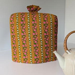 Könnte beinhalten: Ein senffarbener Teewärmer mit vertikalen Streifen aus rosa und weißen Blumenmustern. Der Teewärmer hat eine geraffte Oberseite und befindet sich neben einer weißen Teekanne mit einem geflochtenen Griff.