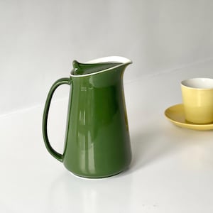 Puede incluir: Una jarra de cerámica verde con borde y asa blancos, junto a una taza y platillo amarillos. La jarra tiene un acabado brillante. La taza y el platillo son un juego a juego. Los artículos están sobre una superficie blanca.
