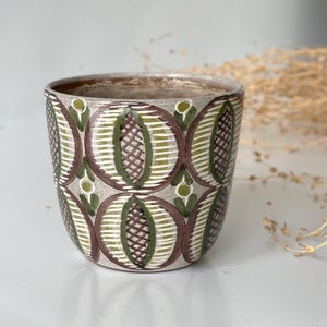 Puede incluir: Una maceta de cerámica con un patrón repetitivo de formas ovaladas marrones, verdes y blancas. La maceta tiene un fondo gris neutro y una superficie ligeramente texturizada. El borde es de color marrón oscuro.
