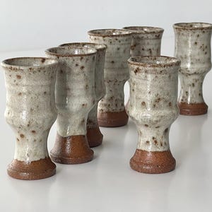 May include: A set of handmade ceramic shot glasses. The glasses feature a speckled, off-white glaze with brown detailing and a reddish-brown base. The design is a unique, handcrafted style.