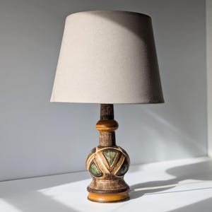 Puede incluir: Una lámpara de mesa de cerámica con un patrón geométrico marrón, verde y amarillo. La lámpara tiene una pantalla de tela beige.