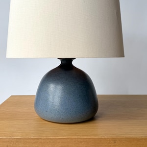 May include: A table lamp featuring a rounded, speckled blue ceramic base with a smooth, matte finish. The lamp is topped with a wide, cylindrical lampshade in a neutral, off-white fabric that provides a soft, diffused light distribution.