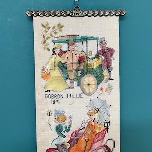 Op de afbeelding: Een vintage naaldkunst wandtapijt met een kleurrijke illustratie van een Gobron-Brillie auto uit 1891. De auto is afgebeeld met vier mensen erin, en de scène speelt zich af tegen een witte achtergrond met een bloemenontwerp.