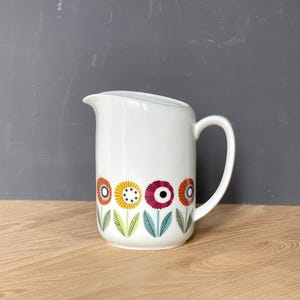 Puede incluir: Jarra de cerámica blanca con asa y boquilla curvadas. La jarra está decorada con una banda de flores estilizadas de colores naranja, amarillo, rosa y turquesa. Colocada sobre una superficie de madera.