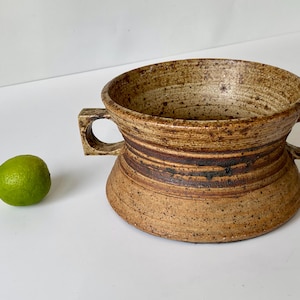 May include: A brown and tan ceramic bowl with two handles. The bowl has a speckled glaze and brown stripes around the rim.