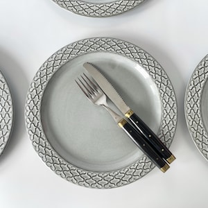 May include: Overhead shot of four gray ceramic plates with a decorative border. A fork and knife with black handles and gold accents rest on one plate. The plates are arranged on a white surface.