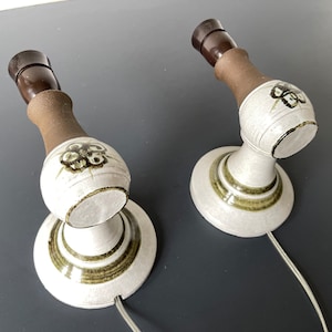 May include: Pair of vintage ceramic wall sconces. Each sconce features a white, bulbous body with a brown neck and a dark brown socket. The white body is decorated with a green and brown floral design and green stripes. Each sconce has a white cord.
