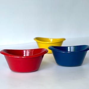 May include: Three small ceramic bowls in red, yellow, and blue. Each bowl has a curved rim and a glossy finish. The bowls are arranged on a white surface. The red bowl is in the foreground.