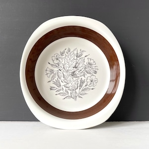 May include: A white ceramic bowl with a brown rim and a black and white floral design. The bowl is ideal for serving snacks or salads.