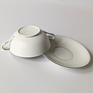 May include: A white porcelain soup bowl with gold trim and a matching saucer. The bowl has a textured design and two handles.