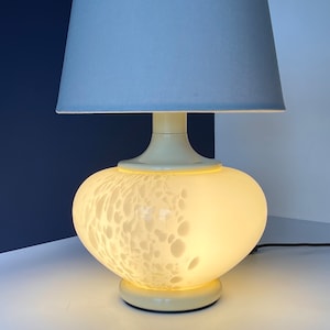 Puede incluir: Una lámpara de mesa blanca con una pantalla de tela gris y una base de vidrio blanco texturizado. La lámpara está encendida y la luz brilla a través del vidrio.