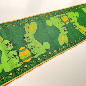 Op de afbeelding: Een groene tafelrunner met een gele rand heeft een herhalend patroon van groene konijnen die gele en groene paaseieren vasthouden. De konijnen hebben gele oren en witte ogen.
