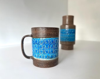 May include: A brown ceramic mug with a blue and white geometric pattern. The mug has a handle and is sitting on a white surface. There is a matching brown ceramic vase in the background.