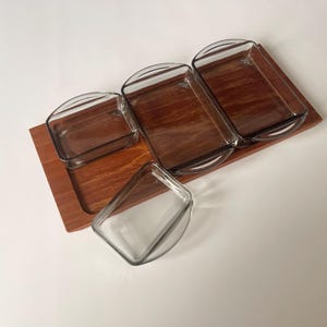 Puede incluir: Un juego de servir con una bandeja rectangular de madera y cuatro recipientes de vidrio transparente. Tres recipientes son rectangulares con asas arqueadas, y uno tiene forma triangular. La madera tiene un tono cálido marrón rojizo.