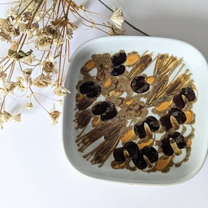 Può includere: Piatto quadrato in ceramica con sfondo bianco e motivo floreale marrone, beige e giallo. Il piatto ha angoli arrotondati e un bordo leggermente rialzato. Fiori secchi nell'angolo in alto a sinistra.