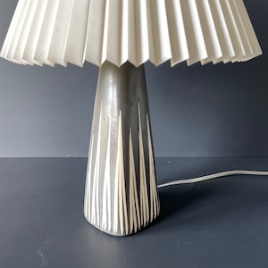 May include: A ceramic table lamp with a white pleated lampshade. The lamp base is gray with white vertical lines.