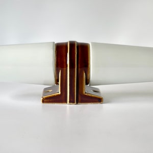 Könnte beinhalten: Ein keramischer Wandhalter für Handtuchstangen mit zwei glatten, weißen, zylindrischen Armen, die horizontal von einer zentralen, dunkelbraun glasierten Basis abstehen. Die Basis hat ein gestuftes, geometrisches Profil mit sichtbaren Schraublöchern.