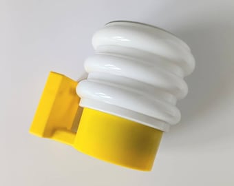 Puede incluir: Una lámpara de pared amarilla y blanca con una pantalla blanca redonda y nervada y una base amarilla. La lámpara tiene un soporte de montaje rectangular amarillo.
