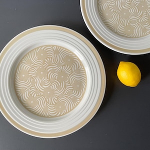 Puede incluir: Dos platos de cerámica blanca con un patrón de espiral beige. Los platos tienen un borde elevado y están apilados uno encima del otro. Un limón amarillo se encuentra sobre una superficie gris oscuro.