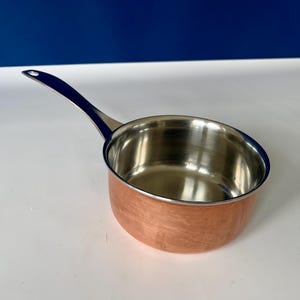 May include: A copper-colored saucepan with a stainless steel interior and a long, black handle. The small cooking pot is designed for heating liquids and sauces. The handle is attached to the side of the pan, allowing for easy lifting and pouring.