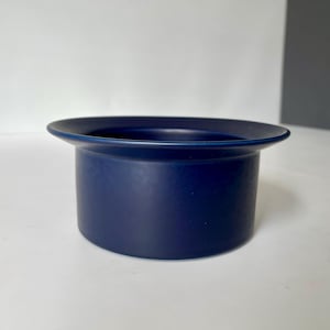 Pode incluir: Um recipiente de cerâmica azul escuro com uma aba larga e plana. O corpo cilíndrico tem um acabamento fosco. O recipiente está sobre uma superfície branca, com um fundo branco e cinza.