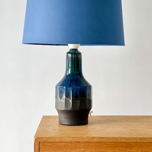 May include: A blue table lamp with a dark blue lampshade. The ceramic base is a gradient of dark blue, teal, and black. The lamp sits on a wooden table.