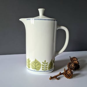Op de afbeelding: Een witte keramische cafetière met een deksel en handvat. De basis heeft een groen en blauw bladeren ontwerp. De cafetière staat op een wit oppervlak met een donkergrijze achtergrond.