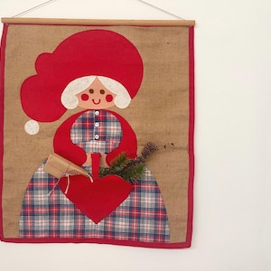 Puede incluir: Un tapiz de pared de arpillera con un diseño de arte popular de una mujer con sombrero y vestido rojos con un estampado de cuadros. Sostiene un corazón rojo y un regalo envuelto. Los bordes están ribeteados en rojo.