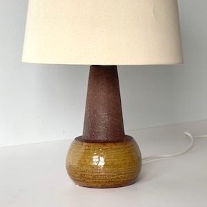May include: A ceramic table lamp with a beige lampshade. The lamp base is a combination of brown and tan tones. The lamp has a textured, cone-shaped body and a rounded base. A white power cord extends from the lamp.