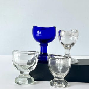 Pode incluir: Quatro taças antigas para lavar os olhos. Uma é azul cobalto profundo, as outras são de vidro transparente. As taças têm formato de tigela arredondada com borda curva e base de pedestal. Estão dispostas sobre uma superfície preta contra um fundo branco.