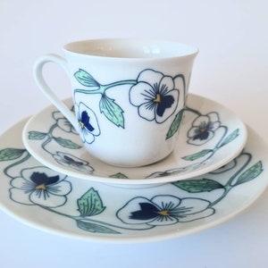 Puede incluir: Un juego de taza y platillo de té de porcelana blanca con un diseño floral que presenta hojas verdes y pensamientos blancos con acentos azules y dorados.