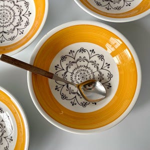 May include: Overhead view of several white bowls with a yellow rim and a black floral pattern. A silver spoon with a wooden handle rests in one bowl. The bowls are on a light grey surface.