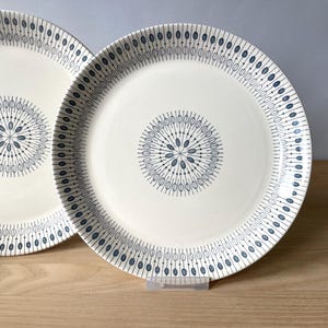 Op de afbeelding: Twee witte keramische borden met een blauw en grijs geometrisch ontwerp. De borden hebben een centraal bloemmotief en een rand van verticale lijnen en ovalen. De borden staan op een houten oppervlak.