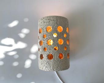 Op de afbeelding: Een cilindrische, getextureerde, grijze wandlamp met talrijke cirkelvormige uitsparingen. De binnenkant gloeit met warm oranje licht. Een wit elektrisch snoer loopt van de onderkant. De lamp is op een witte muur gemonteerd.