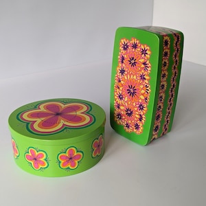 Könnte beinhalten: Zwei grüne Boxen mit Blumendesigns. Die größere Box ist rechteckig und hat ein sich wiederholendes Muster aus orangefarbenen, rosa und violetten Blumen. Die kleinere Box ist rund und hat eine einzelne große orange, rosa und gelbe Blume in der Mitte.