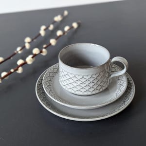 May include: A gray ceramic teacup with a heart pattern sits on a matching saucer. The saucer is on top of a larger, gray saucer with a similar pattern.