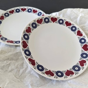 Puede incluir: Dos platos de cerámica blanca con un diseño de borde azul y rojo. El borde presenta un patrón repetitivo de corazones y círculos.