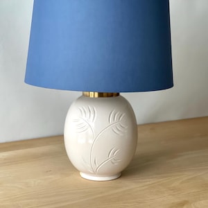 May include: A cream-colored ceramic table lamp with a blue lampshade. The lamp base has a gold-colored band and a floral leaf design. The lamp is on a wooden surface.
