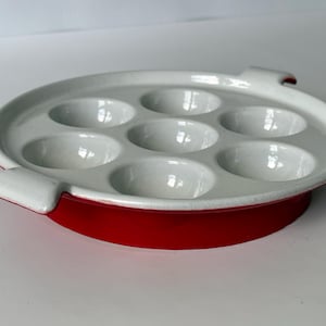 Może przedstawiać: Okrągłe, białe naczynie ceramiczne do ślimaków z sześcioma okrągłymi wgłębieniami. Zewnętrzna część naczynia ma żywy czerwony kolor i posiada dwa małe, zintegrowane uchwyty po przeciwnych stronach, ułatwiające chwytanie i przenoszenie.