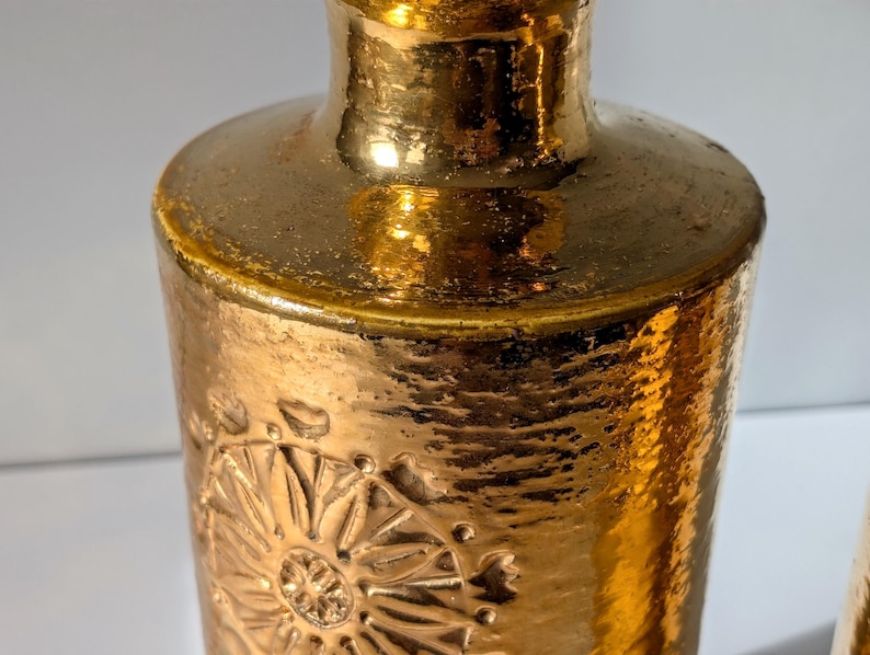 May include: A pair of gold ceramic vases with a textured surface and embossed floral designs. The vases have a slightly flared top and a rounded bottom.
