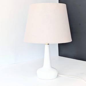 Puede incluir: Una lámpara de mesa de cerámica blanca con una pantalla de tela blanca. La lámpara tiene un diseño simple y moderno.