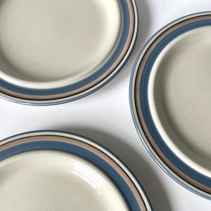 Könnte beinhalten: Drei cremefarbene Keramikplatten mit blau-braunem Streifenrand. Die Teller sind auf einer weißen Oberfläche angeordnet und präsentieren ihr schlichtes, aber elegantes Design. Ideal zum Servieren von Mahlzeiten.